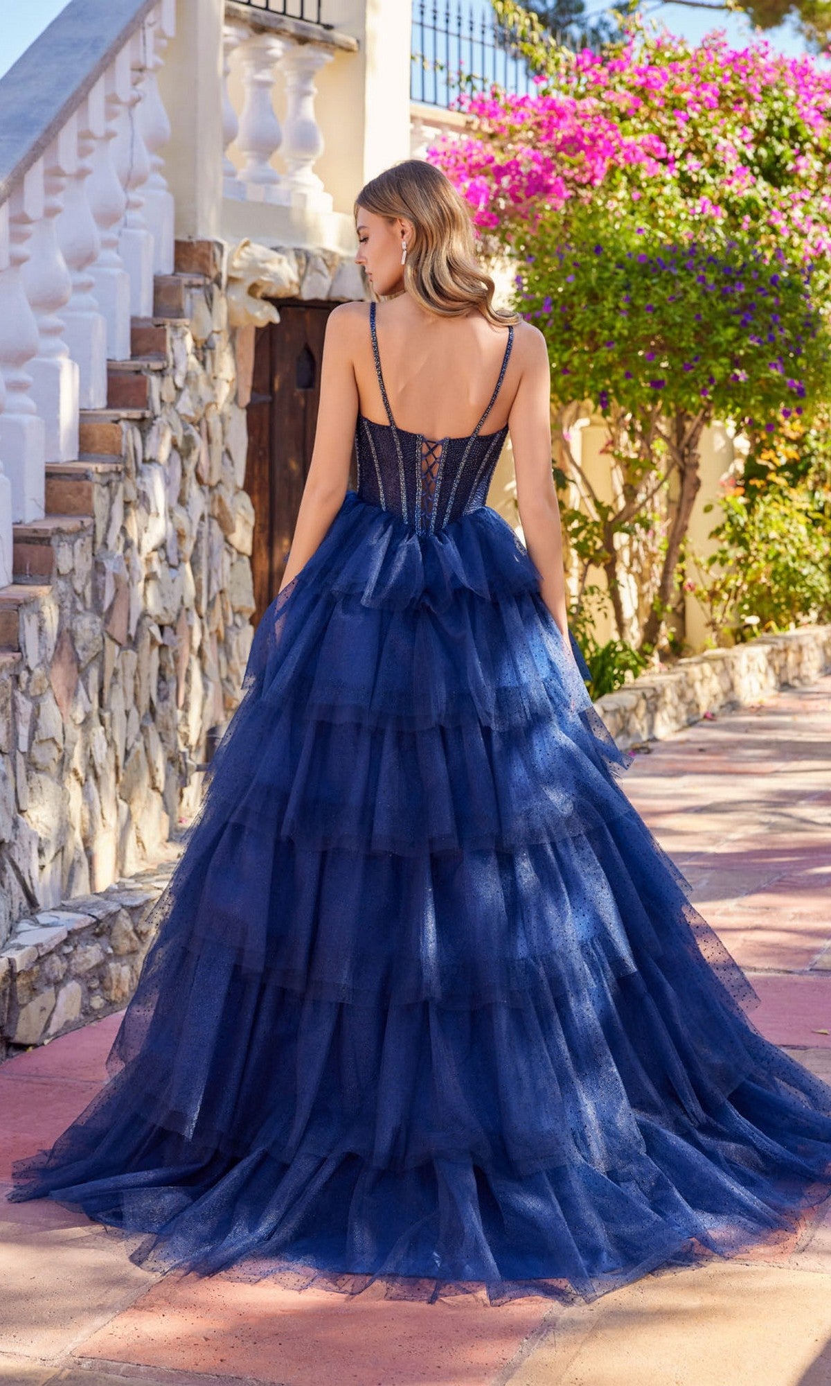 A woman stands outdoors before stone stairs, wearing the Juliet JT2611N long prom dress—a navy blue glitter tulle ball gown with a corset-style back and tiered skirt.
